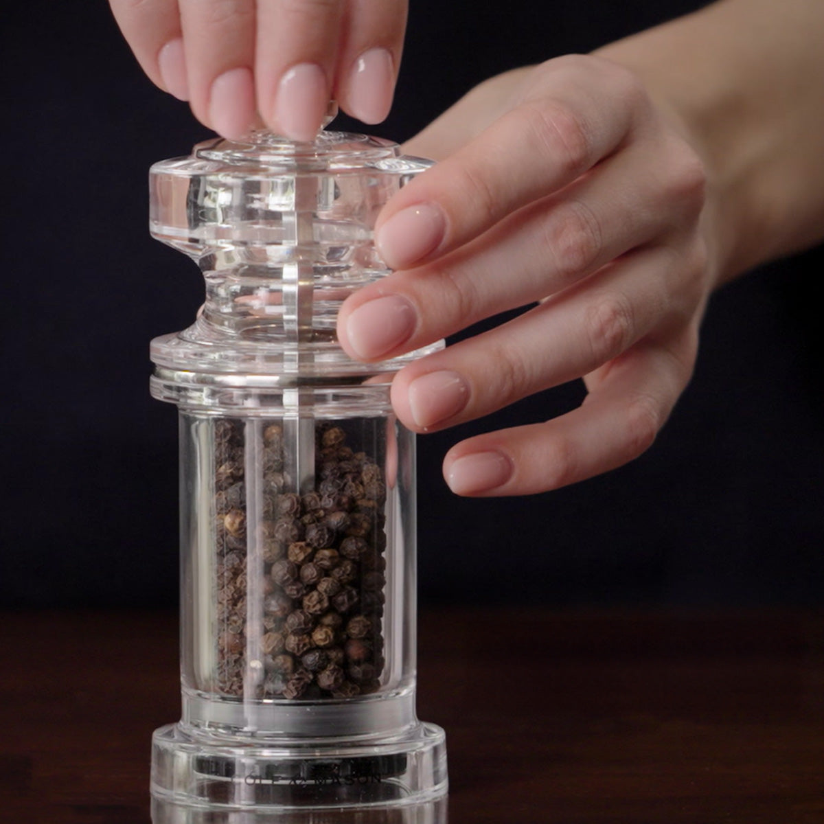 A person’s hands are shown grinding black peppercorns with a clear, acrylic pepper mill against a dark background.