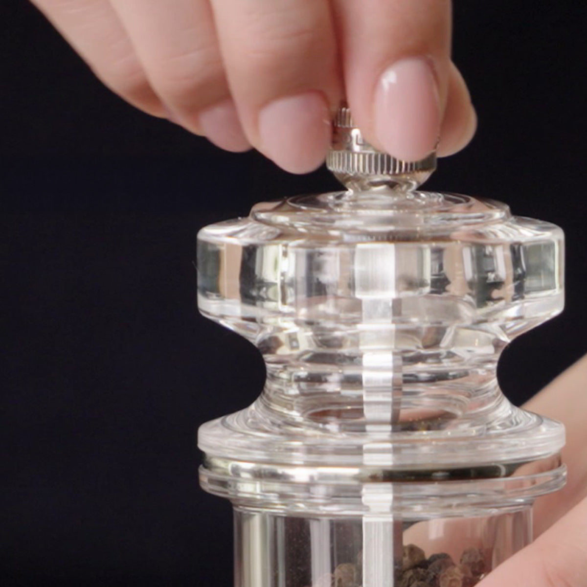 A close-up of a hand adjusting the top knob of a clear acrylic pepper grinder, with another hand holding the base.