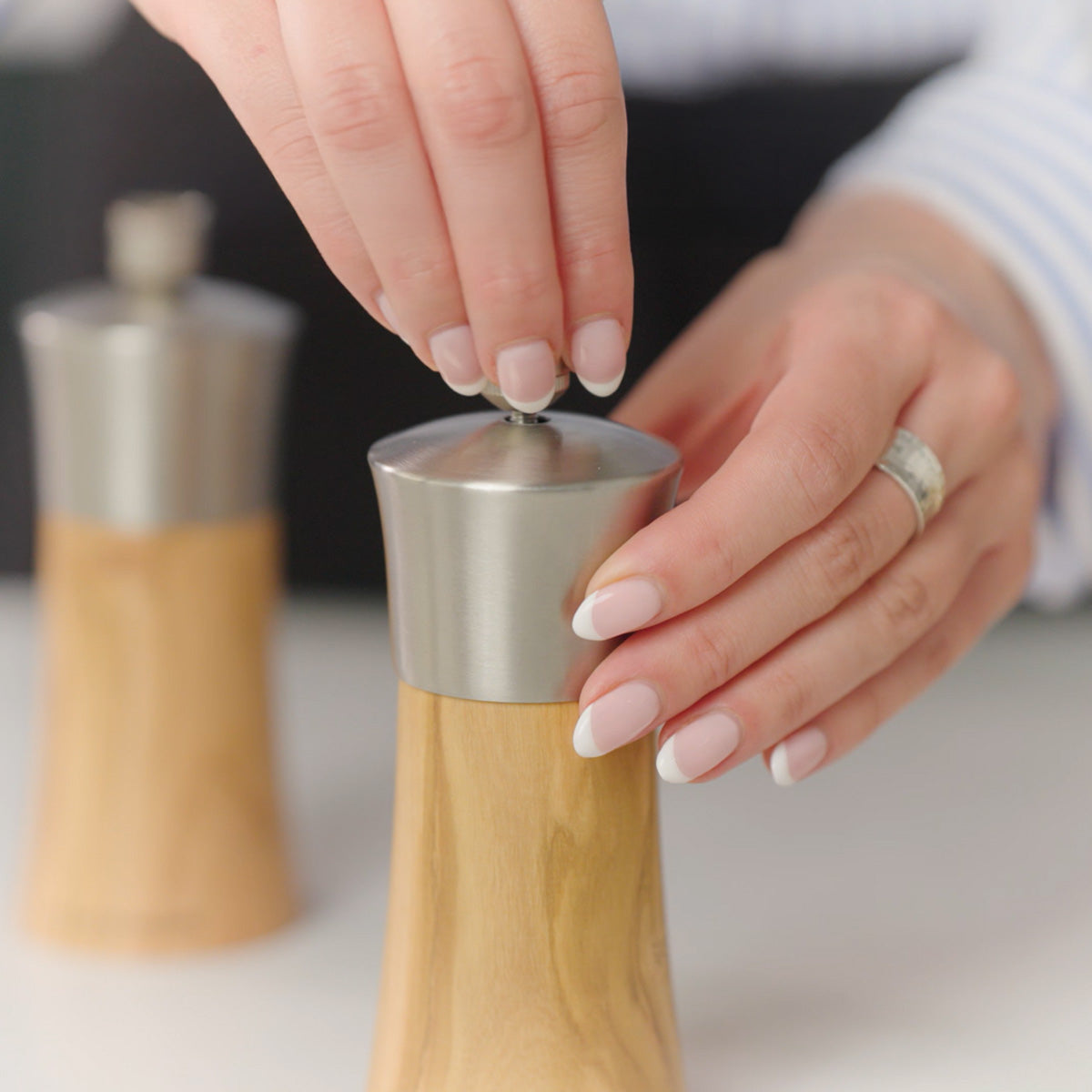 A person with French-manicured nails and a silver ring adjusting the top of a wooden pepper grinder, with another similar grinder blurred in the background.