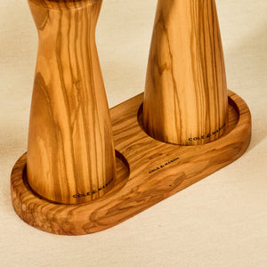 Two Cole & Mason wooden salt and pepper mills stand upright in a Portobello Olive Wood Mill Tray, resting on a beige fabric surface.