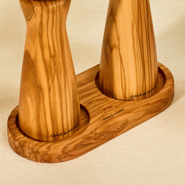 Two Cole & Mason wooden salt and pepper mills stand upright in a Portobello Olive Wood Mill Tray, resting on a beige fabric surface.