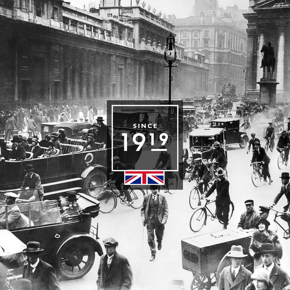 Black and white photo of a busy London street in 1919, showing people walking, riding bicycles, and driving vintage cars, with old buildings and a Union Jack flag overlayed at the center. Text reads Since 1919.