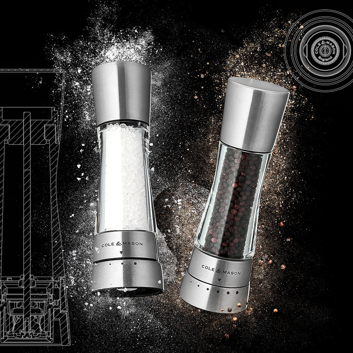 Two Cole & Mason grinders tilted diagonally, one filled with salt crystals and the other with black peppercorns, set against a dark background with dramatic bursts of salt and pepper particles.
