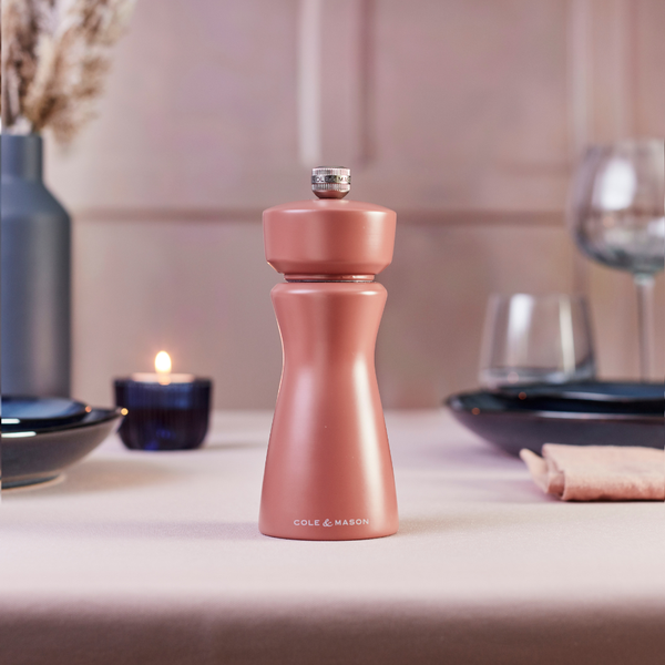 A Cole & Mason Kenton Salt & Pepper Mill in Cinnamon rests on a beige table, surrounded by plates, glasses, a napkin, and a lit candle in a cozy dining setting—an elegant addition to any modern kitchen with its durable ceramic grinder.