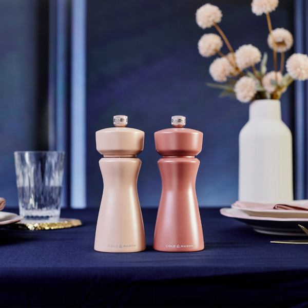 Two stylish Cole & Mason Kenton Salt & Pepper Mills in Cinnamon, featuring durable ceramic grinders, sit on a dark blue tablecloth, complemented by elegant table settings and a white vase of dried flowers in the background—perfect for adding a touch of sophistication to any modern kitchen.