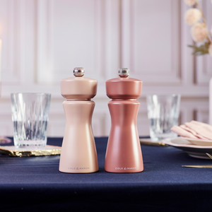 Two metallic Cole & Mason Kenton Salt & Pepper Mills in Himalayan Salt sit on a navy blue tablecloth, featuring a high-quality ceramic grinder perfect for freshly cracking pink Himalayan salt or peppercorns. Glassware, plates, and hints of a blurred hand and flowers in the background enhance the elegant and formal setting.