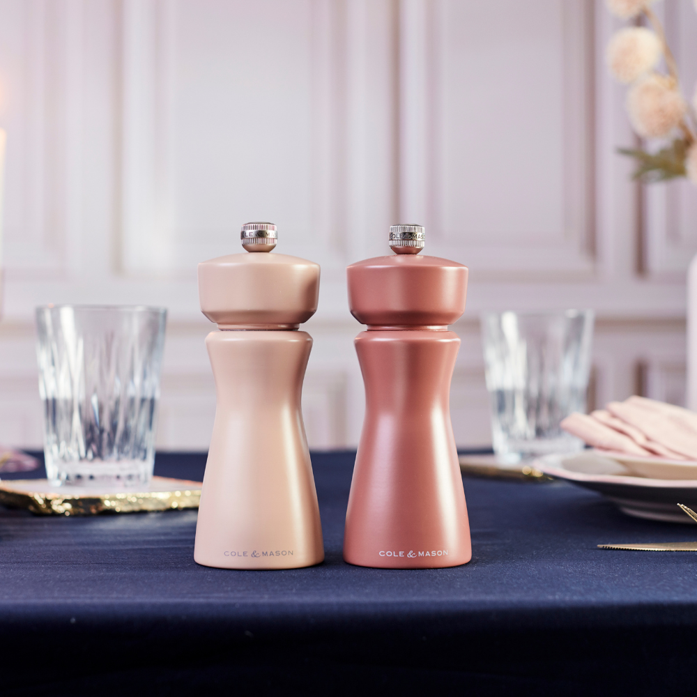 Two metallic Cole & Mason Kenton Salt & Pepper Mills in Himalayan Salt sit on a navy blue tablecloth, featuring a high-quality ceramic grinder perfect for freshly cracking pink Himalayan salt or peppercorns. Glassware, plates, and hints of a blurred hand and flowers in the background enhance the elegant and formal setting.