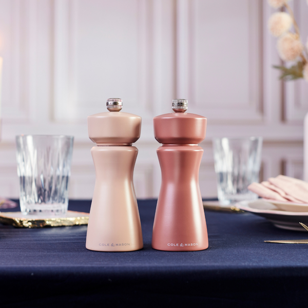 Two metallic Cole & Mason Kenton Salt & Pepper Mills in Himalayan Salt sit on a navy blue tablecloth, featuring a high-quality ceramic grinder perfect for freshly cracking pink Himalayan salt or peppercorns. Glassware, plates, and hints of a blurred hand and flowers in the background enhance the elegant and formal setting.