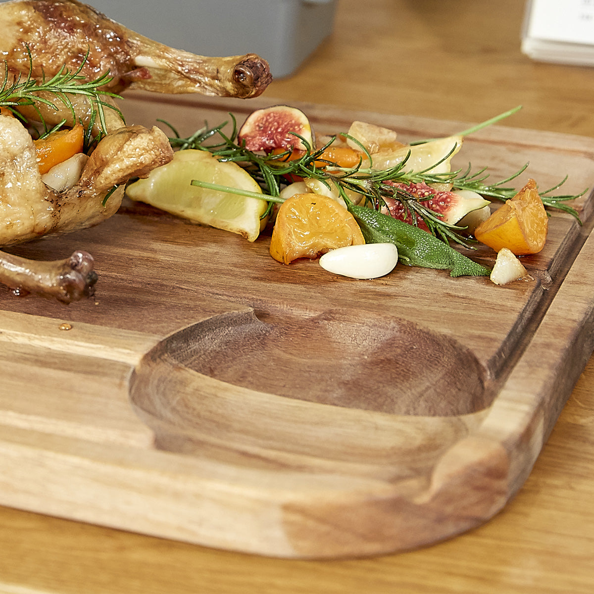 A wooden cutting board holding part of a roasted chicken garnished with rosemary, garlic cloves, figs, and sliced yellow tomatoes on a wooden table.