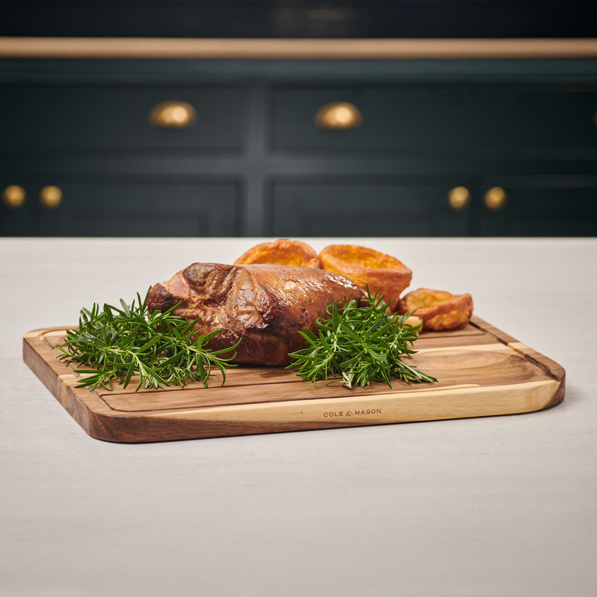 A roast joint of meat with crispy potatoes and fresh sprigs of rosemary is arranged on a wooden cutting board atop a light-colored countertop, with dark cabinets and golden handles in the background.