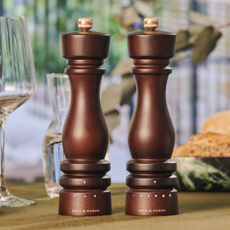 Two dark wooden salt and pepper grinders labeled Cole & Mason stand side by side on a table, with glassware, a loaf of bread, and blurred greenery in the background.