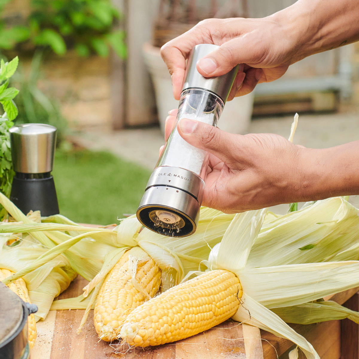 A person seasons fresh corn on the cob with a silver salt grinder on a wooden surface outdoors. Unhusked corn and green herbs are visible in the background.