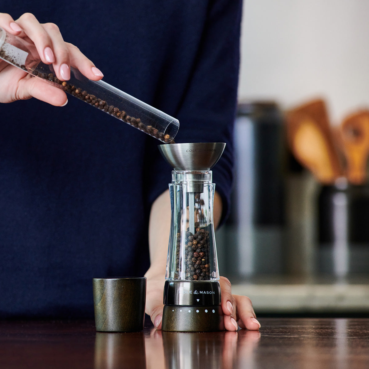 A person pours black peppercorns from a glass tube into the top of a transparent pepper grinder on a kitchen counter, with utensils blurred in the background.