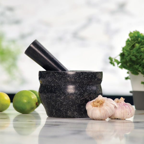A black mortar and pestle sits on a marble countertop next to two garlic bulbs, two limes, and a bunch of fresh green herbs in the background.