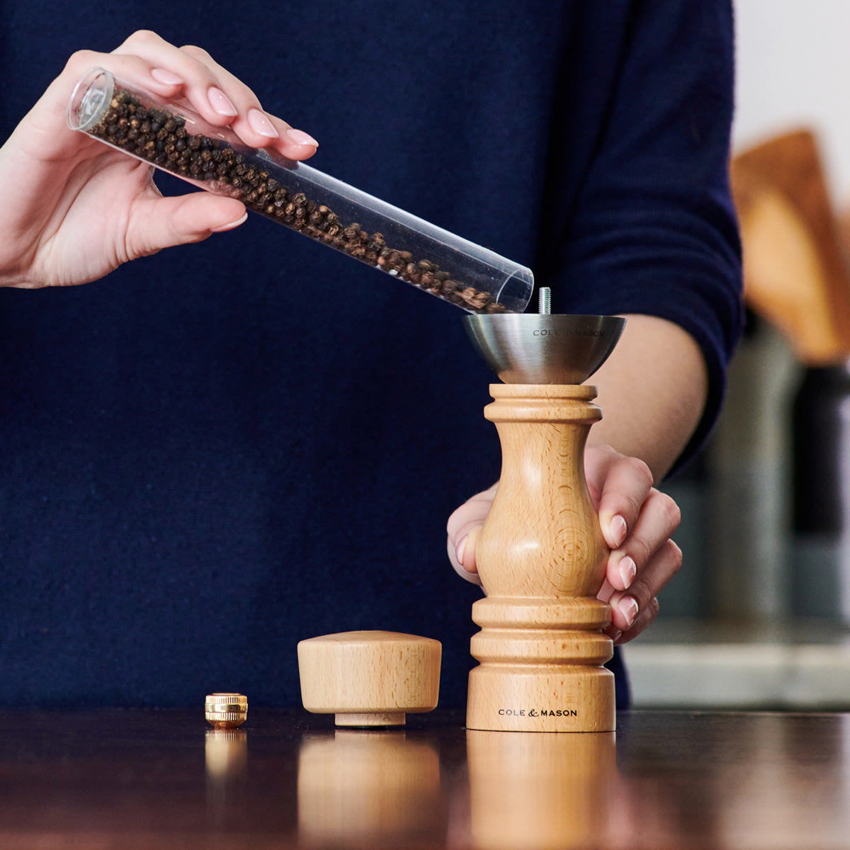 A person pours whole peppercorns from a glass tube into a wooden pepper grinder with a metal funnel. The grinder’s cap and adjuster are on the table nearby.