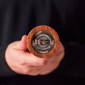 #pepper A person holds a Cole & Mason Portobello Olive Wood Salt & Pepper Mill horizontally, showing its adjustable grind metal mechanism and branded base against a dark, blurred background.