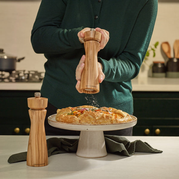 #salt&pepperset A person in a green sweater grinds pepper over a savory pie using the Cole & Mason Portobello Olive Wood Salt & Pepper Mill, with its precision mechanism. Another mill and kitchen utensils appear in the background.