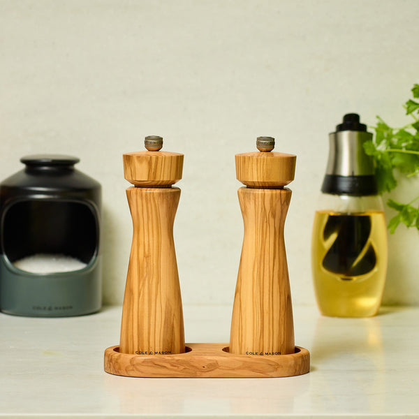Two wooden mills sit on the Cole & Mason Portobello Olive Wood Mill Tray atop a kitchen counter, with a black salt cellar, oil bottle, and fresh green herbs behind them for a stylish countertop organizer.