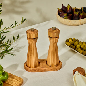 The Cole & Mason Portobello Olive Wood Mill Tray holds two wooden salt and pepper grinders on a white tablecloth, accompanied by green olives, figs in a bowl, a tomato, and olive branches.