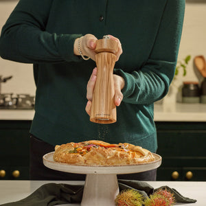 #pepper A person in a green sweater grinds pepper with Cole & Mason Portobello Olive Wood Salt & Pepper Mills over a veggie-topped savory pie on a cake stand, with rambutan fruits nearby.