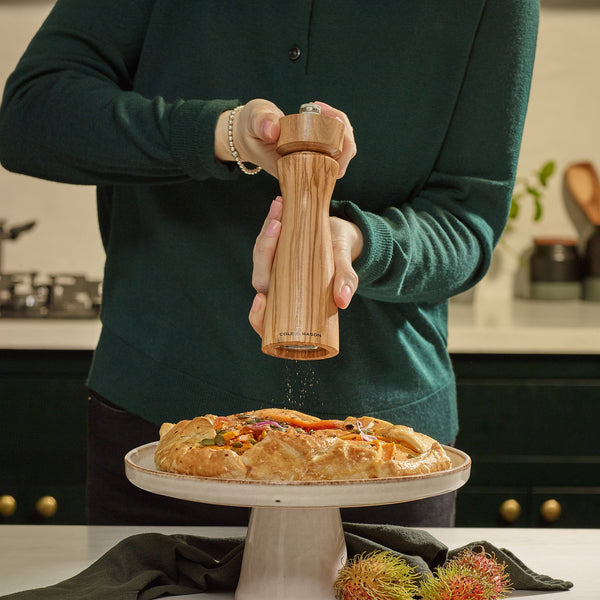 #pepper A person in a green sweater grinds pepper with Cole & Mason Portobello Olive Wood Salt & Pepper Mills over a veggie-topped savory pie on a cake stand, with rambutan fruits nearby.