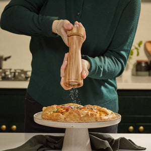 #salt In a modern kitchen, someone in a green sweater grinds pepper with Cole & Mason Portobello Olive Wood Salt & Pepper Mills over a rustic pie on a stand.