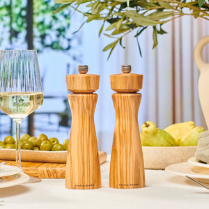 #salt&pepperset The Cole & Mason Portobello Olive Wood Salt & Pepper Mills are displayed on a set dining table with white wine, green olives, a plate, and fruit, surrounded by greenery. Their precision grinding mechanism ensures perfect seasoning.