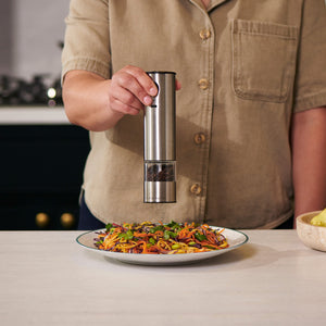 A person in a beige shirt uses the Cole & Mason Battersea USB Electric Salt & Pepper Mills to season a colorful vegetable pasta on a white kitchen counter.