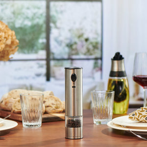 The Cole & Mason Battersea USB Electric Salt & Pepper Mills, featuring a ceramic grinder and stainless steel design, sit on a set dining table with glassware, bread, oil, and red wine—perfect for stylish one-handed use during meals.