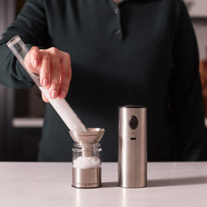 A person pours coarse salt into a jar using a funnel. Beside the jar is the Cole & Mason Battersea USB Electric Salt & Pepper Mill, featuring one-handed operation, on a white countertop.