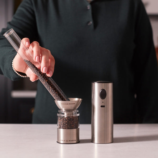 A person pours whole black peppercorns into a small grinder, with the Cole & Mason Battersea USB Electric Salt & Pepper Mills—designed for one-handed use—next to it on a white countertop.
