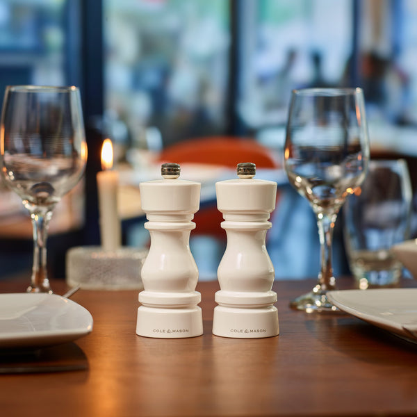 #salt&pepperset The Cole & Mason London White Acrylic Salt & Pepper Mills 150mm, featuring adjustable grinding, are displayed on a dining table with wine glasses, a candle, and plates in a softly blurred restaurant setting.