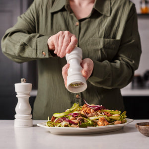 #salt A person in a green shirt seasons roasted vegetables with the Cole & Mason London White Acrylic Salt & Pepper Mills 150mm, featuring an adjustable grinding mechanism, while another high-gloss mill rests on the countertop nearby.