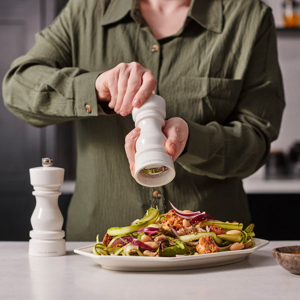 #salt A person in a green shirt seasons roasted vegetables with the Cole & Mason London White Acrylic Salt & Pepper Mills 150mm, featuring an adjustable grinding mechanism, while another high-gloss mill rests on the countertop nearby.