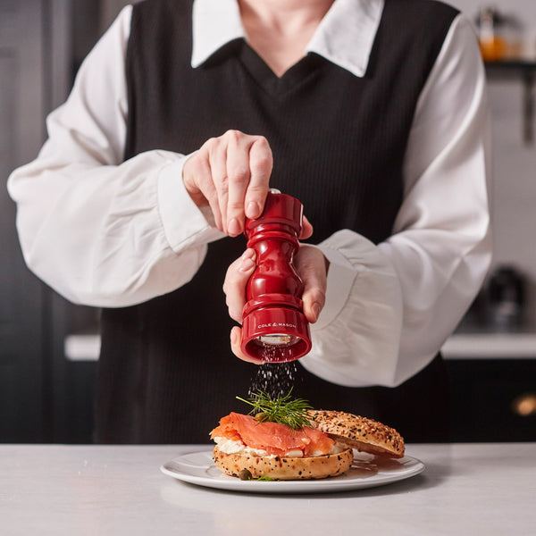 #salt Wearing a white blouse and black vest, someone grinds pepper from a Cole & Mason London Red Acrylic Salt & Pepper Mill (150mm) onto a smoked salmon and dill bagel served on a white plate.