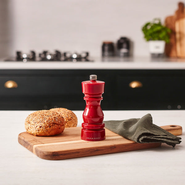 #salt The Cole & Mason London Red Acrylic Salt & Pepper Mills 150mm, featuring an adjustable ceramic grinder, rest on a wooden board beside two seeded rolls and a green napkin, with a stove and potted plant in the kitchen background.