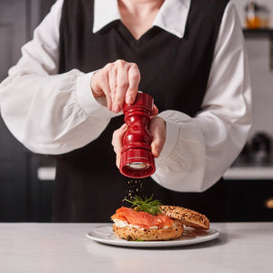#pepper A person in a black vest over a white shirt grinds pepper onto smoked salmon and dill bagel using the Cole & Mason London Red Acrylic Salt & Pepper Mill 150mm, with the bagel on a white plate on the kitchen counter.