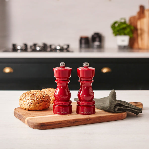 #salt&pepperset Two Cole & Mason London Red Acrylic Salt & Pepper Mills 150mm with adjustable ceramic grinders sit on a wooden cutting board next to seeded bread rolls and a dark napkin, with a stove and green cabinet in the background.