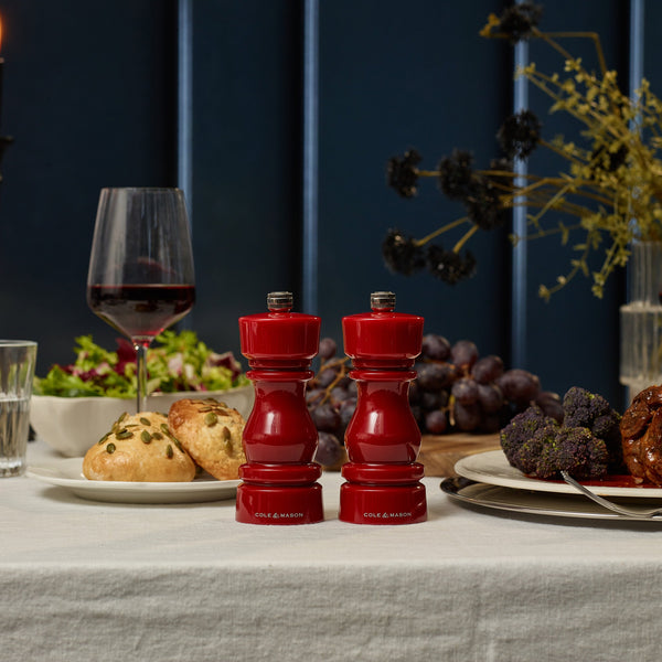 #salt&pepperset A dining table set with two Cole & Mason London Red Acrylic Salt & Pepper Mills 150mm with adjustable ceramic grinders, a glass of red wine, salad, bread rolls, grapes, broccoli, and meat on a white tablecloth against a dark background.