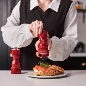 #salt&pepperset A person in a white blouse and black vest uses a Cole & Mason London Red Acrylic Salt & Pepper Mill 150mm to grind pepper onto a bagel with salmon and dill. Another matching mill sits on the table beside the plate.