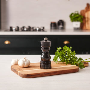 The Cole & Mason London Black Acrylic Salt & Pepper Mill 150mm rests on a wooden cutting board with parsley and garlic, while kitchen items and a stove appear blurred in the background.