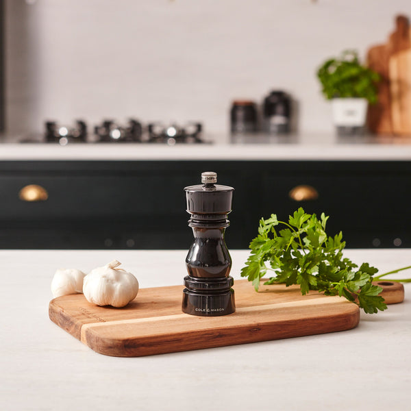 The Cole & Mason London Black Acrylic Salt & Pepper Mill 150mm rests on a wooden cutting board with parsley and garlic, while kitchen items and a stove appear blurred in the background.