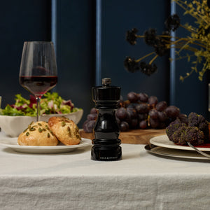The Cole & Mason London Black Acrylic Salt & Pepper Mills 150mm sit on a set table with red wine, bread rolls, salad, cheese balls, and grapes atop a white tablecloth.