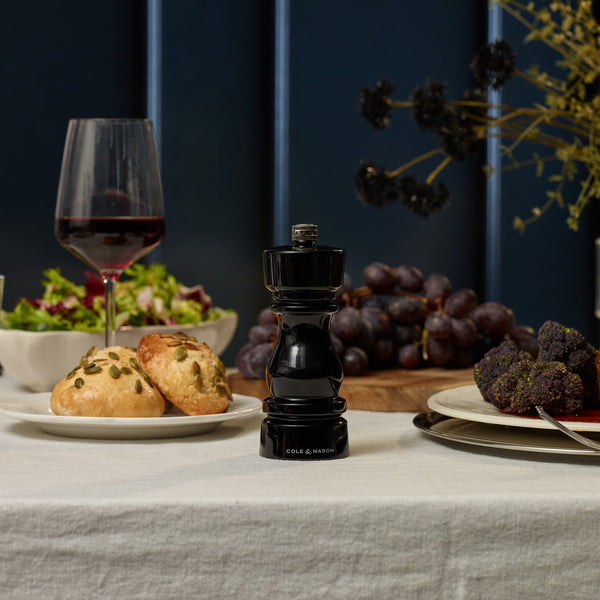 The Cole & Mason London Black Acrylic Salt & Pepper Mills 150mm sit on a set table with red wine, bread rolls, salad, cheese balls, and grapes atop a white tablecloth.