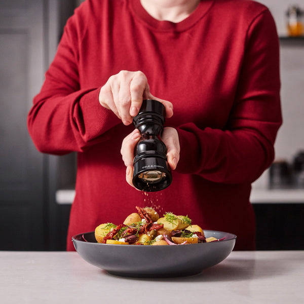 Wearing a red sweater, a person uses Cole & Mason London Black Acrylic Salt & Pepper Mills 150mm to grind pepper over a bowl with potatoes, herbs, and other ingredients on the kitchen counter.