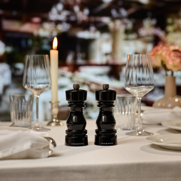 The Cole & Mason London Black Acrylic Salt & Pepper Mills 150mm are set on a white tablecloth amid wine glasses, a candle, plates, napkins, and flowers for an elegant dining experience.