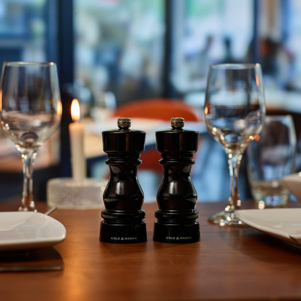 Two Cole & Mason London Black Acrylic Salt & Pepper Mills 150mm with adjustable grind settings sit on a dinner table with plates, empty wine glasses, and a lit candle, creating an elegant and inviting atmosphere.