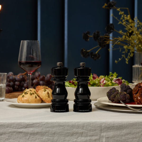 A dining table set with salad, bread, grapes, broccoli, and meat features Cole & Mason London Black Acrylic Salt & Pepper Mills 150mm with adjustable grind settings, a glass of red wine, and a floral arrangement in the background.