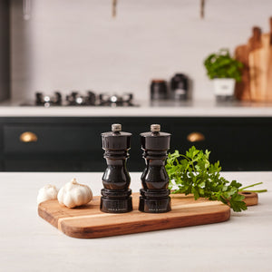 The Cole & Mason London Black Acrylic Salt & Pepper Mills 150mm, featuring adjustable grind settings, rest on a wooden board beside garlic and parsley on a white countertop in a modern kitchen.