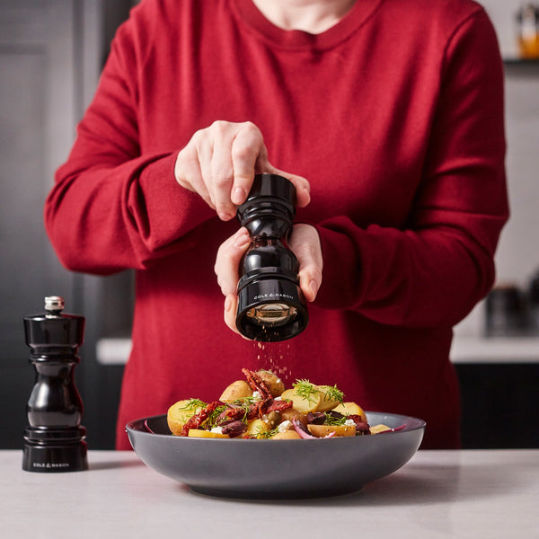 A person in a red sweater grinds black pepper over potatoes and herbs using Cole & Mason London Black Acrylic Salt & Pepper Mills 150mm, featuring adjustable grind settings. Another mill sits beside the bowl.